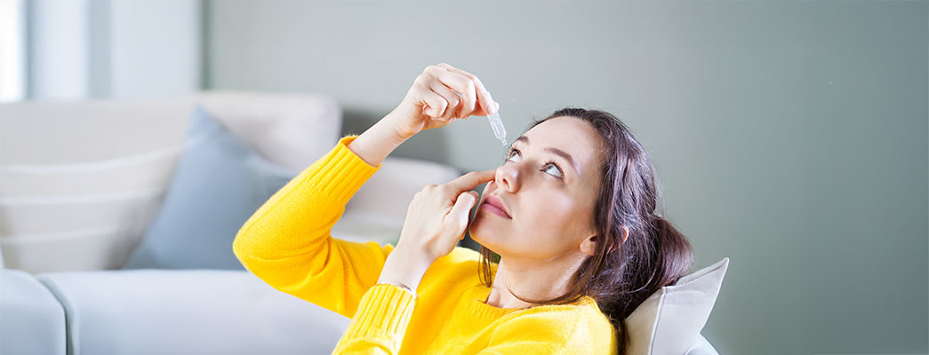 Junge Frau lehnt sich auf dem Sofa nach hinten und tropft sich Augentropfen ins Auge