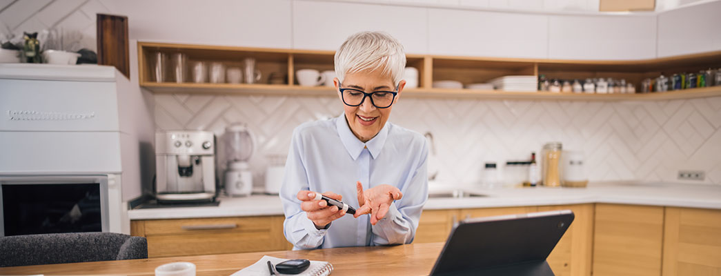 Ältere Frau sitzt in ihrer Küche und testet sich mit einem Messgerät auf Diabetes