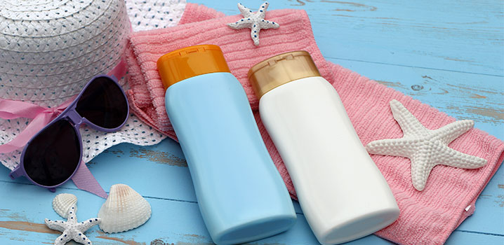 Zwei Tuben Sonnenmilch auf blauem Holzhintergrund mit Handtüchern, Sonnenbrille und Strohhut
